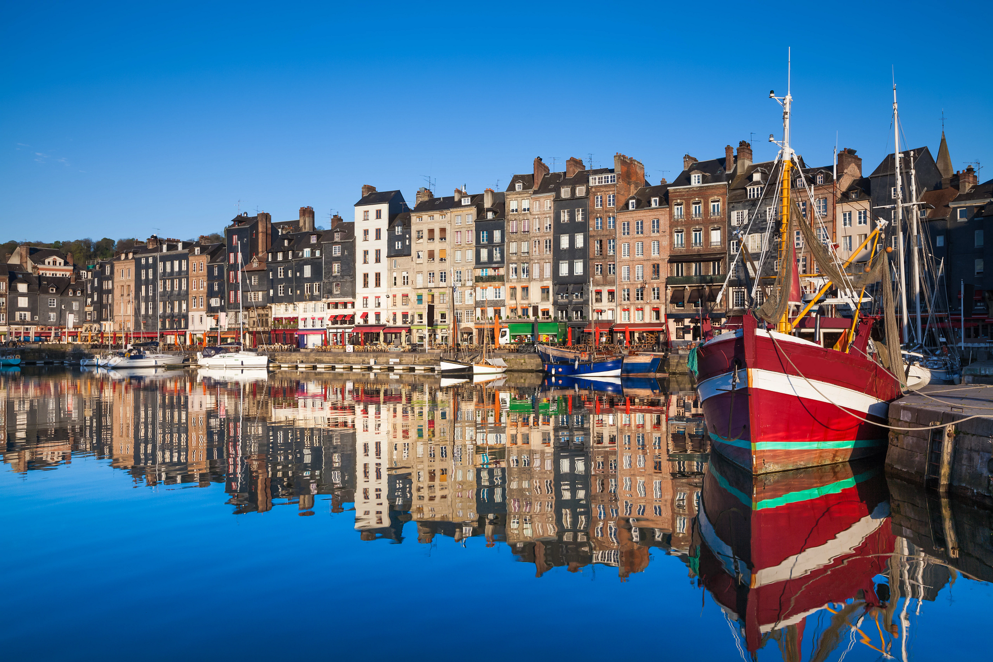 Colorful buildings and a red fishing boat reflected in the calm waters of Honfleur Harbor, France, under a clear blue sky.