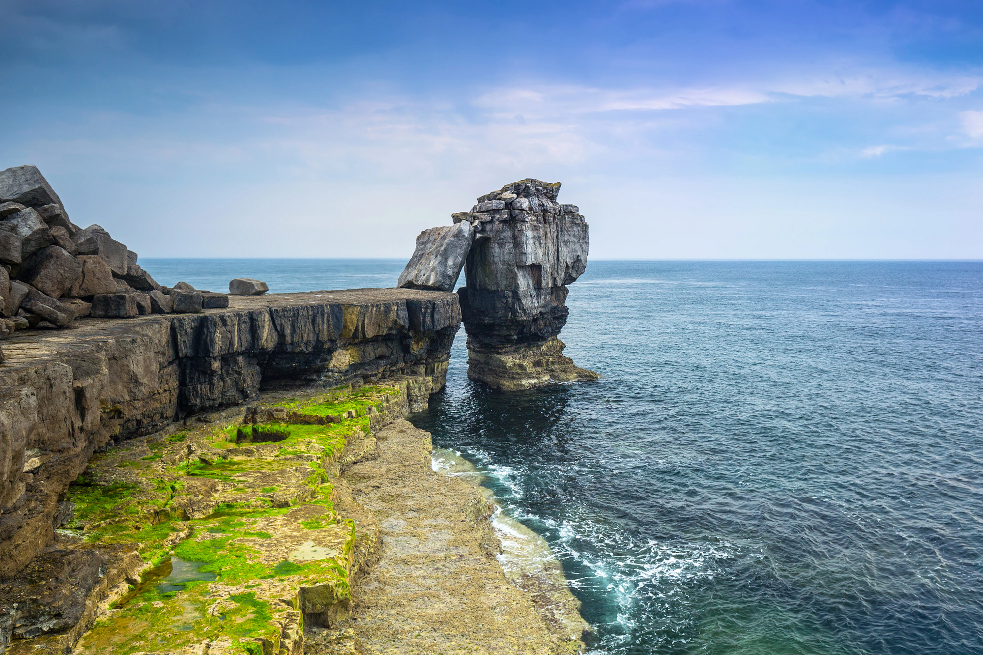 A dramatic shot of Pulpit Rock, a natural rock arch, against a serene blue ocean and sky in Portland, England.