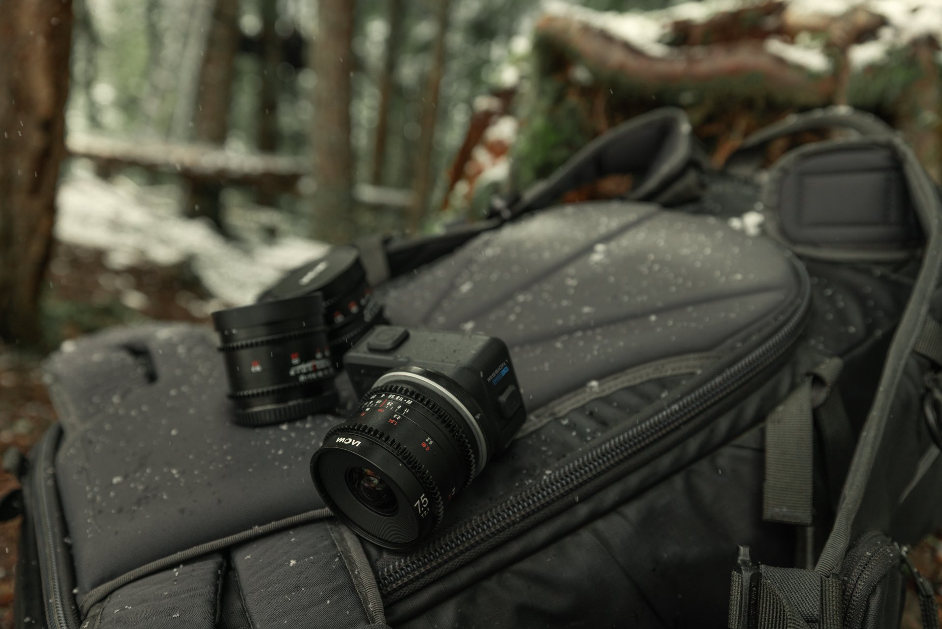 Mission 1 camera and lenses sitting on a black backpack in a snowy forest.