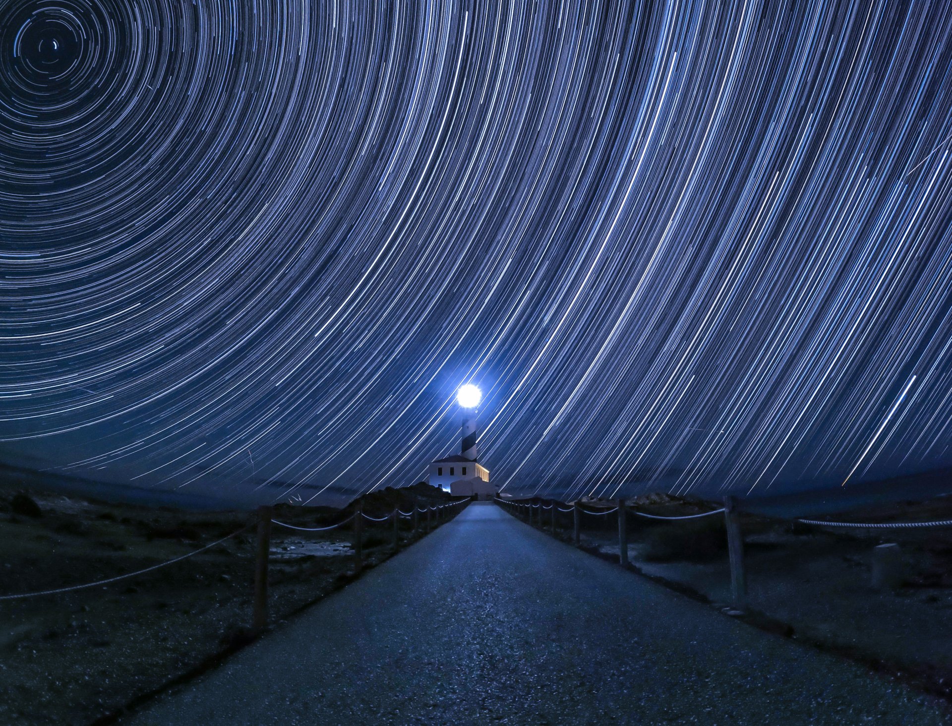 A lighthouse illuminated under a vibrant star trail night sky.
