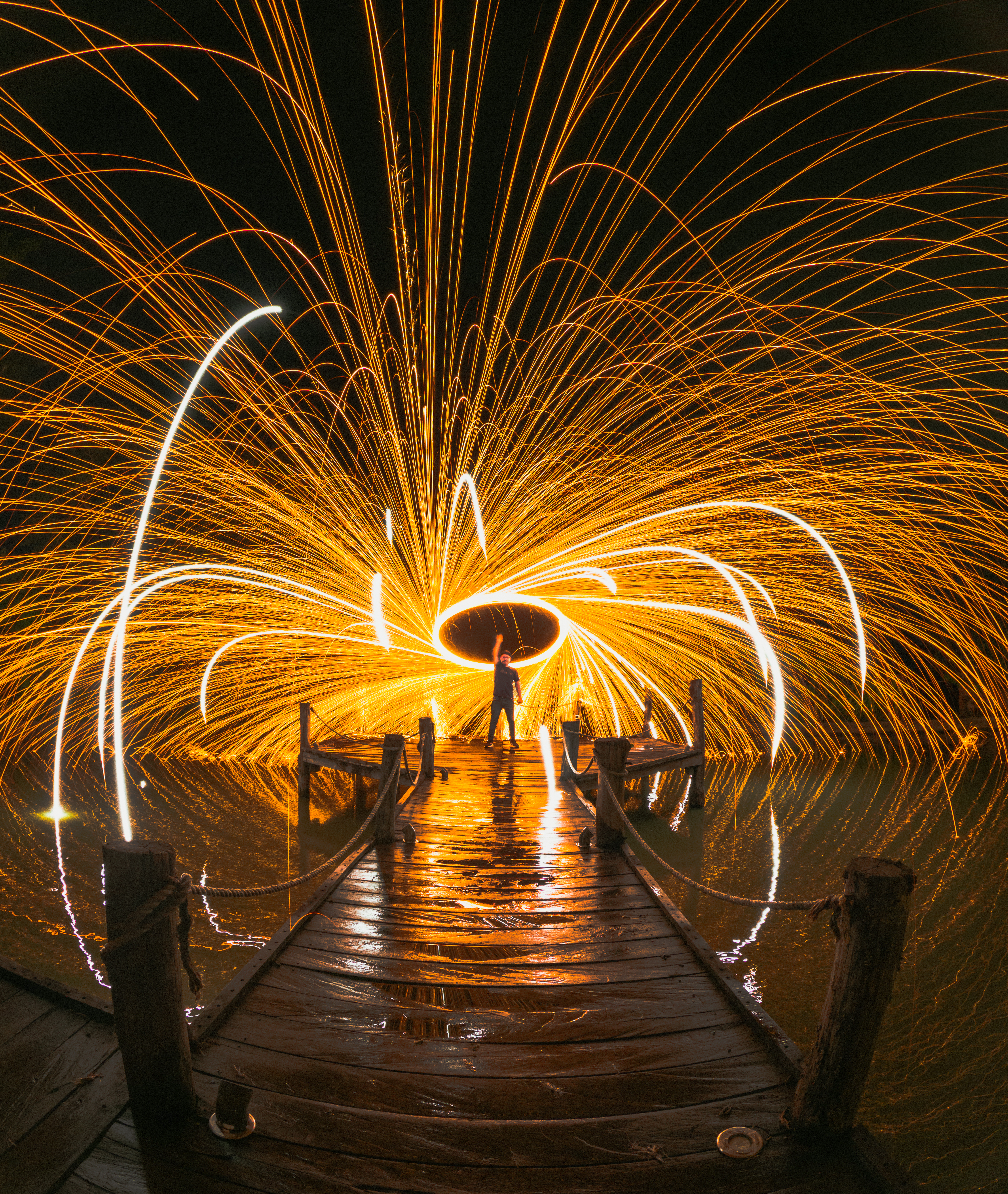 A person swinging burning steel wool to create a circular light display on a wooden pier at night.